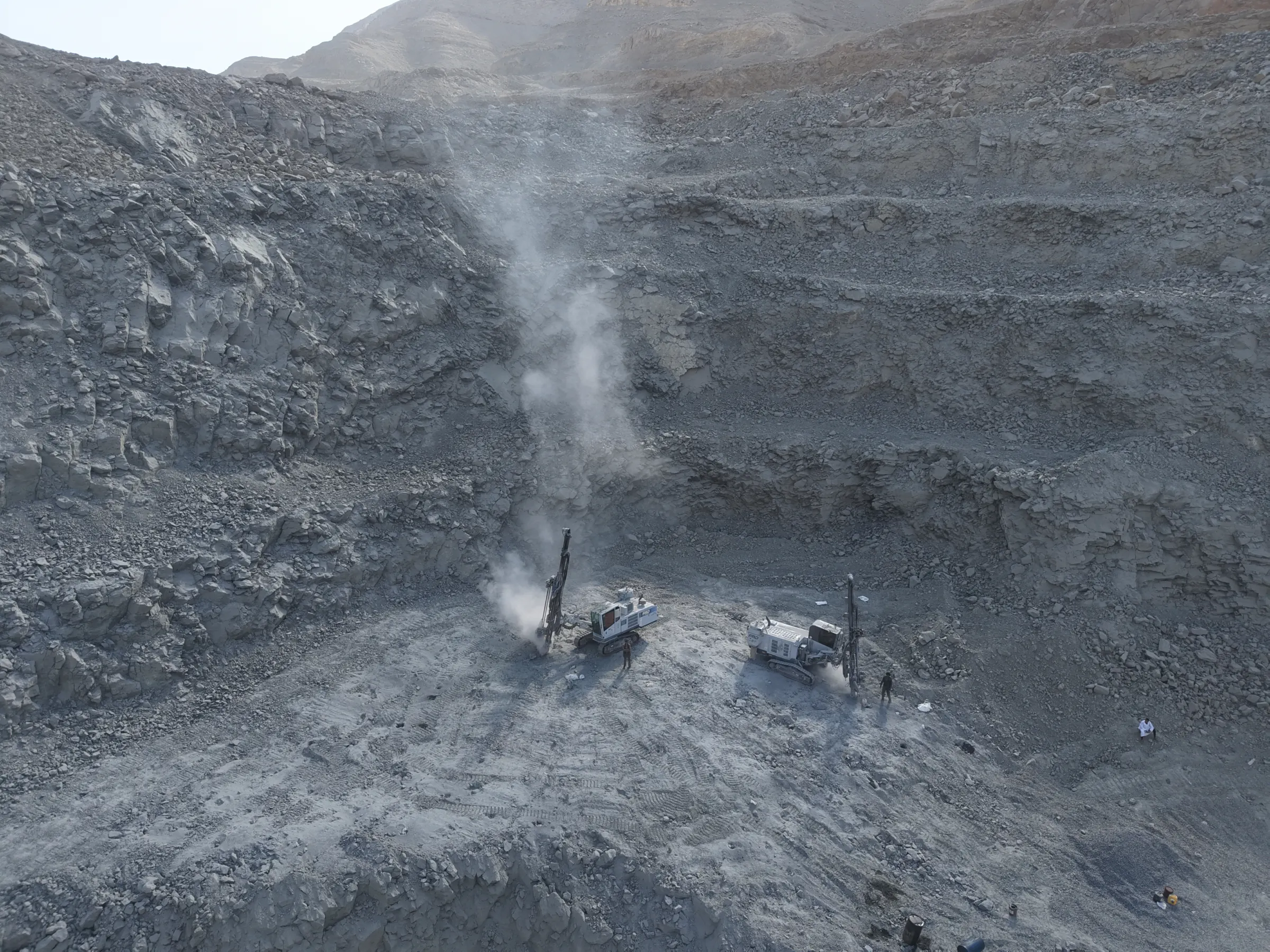 Drill rigs working on a benched quarry face, dust rising from the working floor